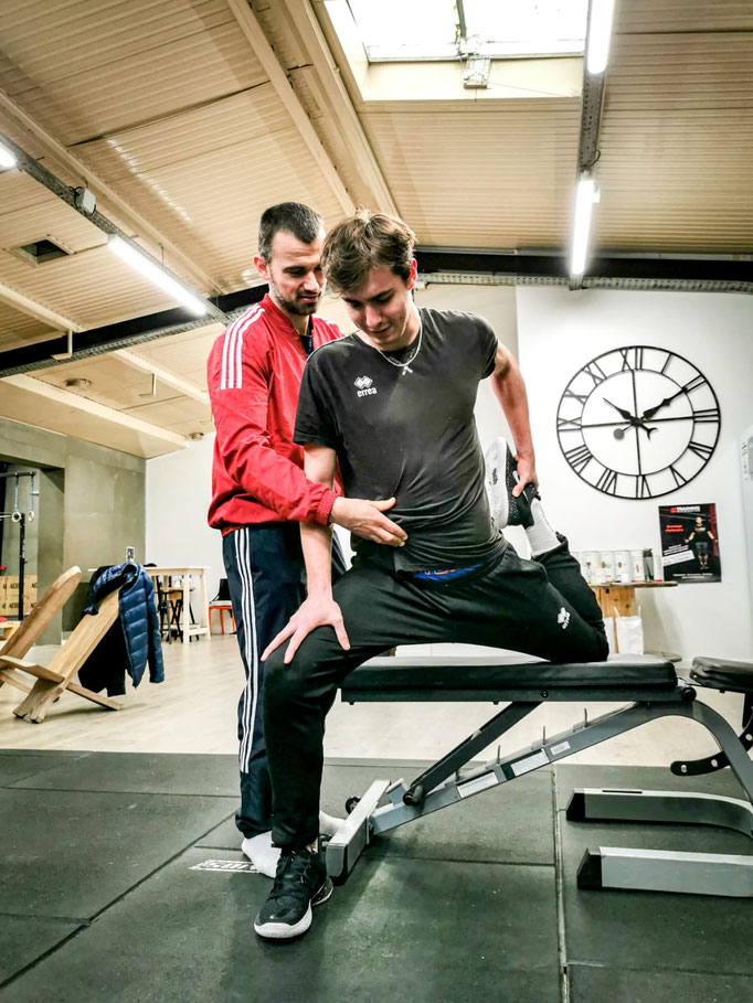 Préparateur physique accompagnant un jeune sportif dans un étirement des fléchisseurs de hanche sur banc de musculation
